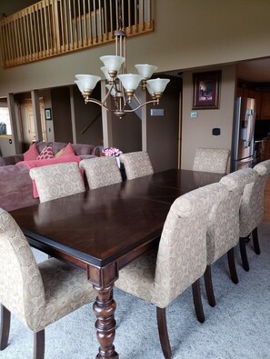 A dining area with a large wooden table surrounded by patterned chairs, a chandelier, and a view of a kitchen with a refrigerator.