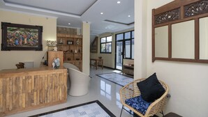 A reception area with a wooden reception desk, a wicker chair with a black cushion, a large framed painting, and a view of the adjacent room through a glass door.