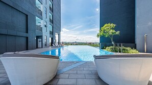A modern outdoor pool area with two wicker lounge chairs, a clear blue sky, and a view of a cityscape in the background. - Dream City By Himmel (Seri Kembangan)