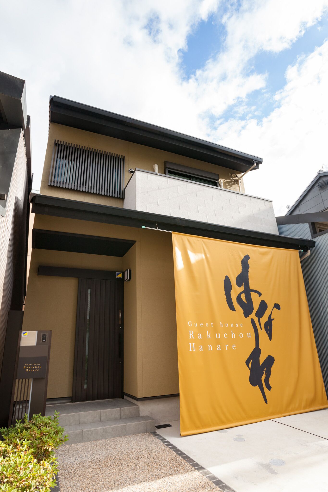 A modern building with a yellow banner displaying Japanese characters and the text "Guest house Rakuuchou Hanare".