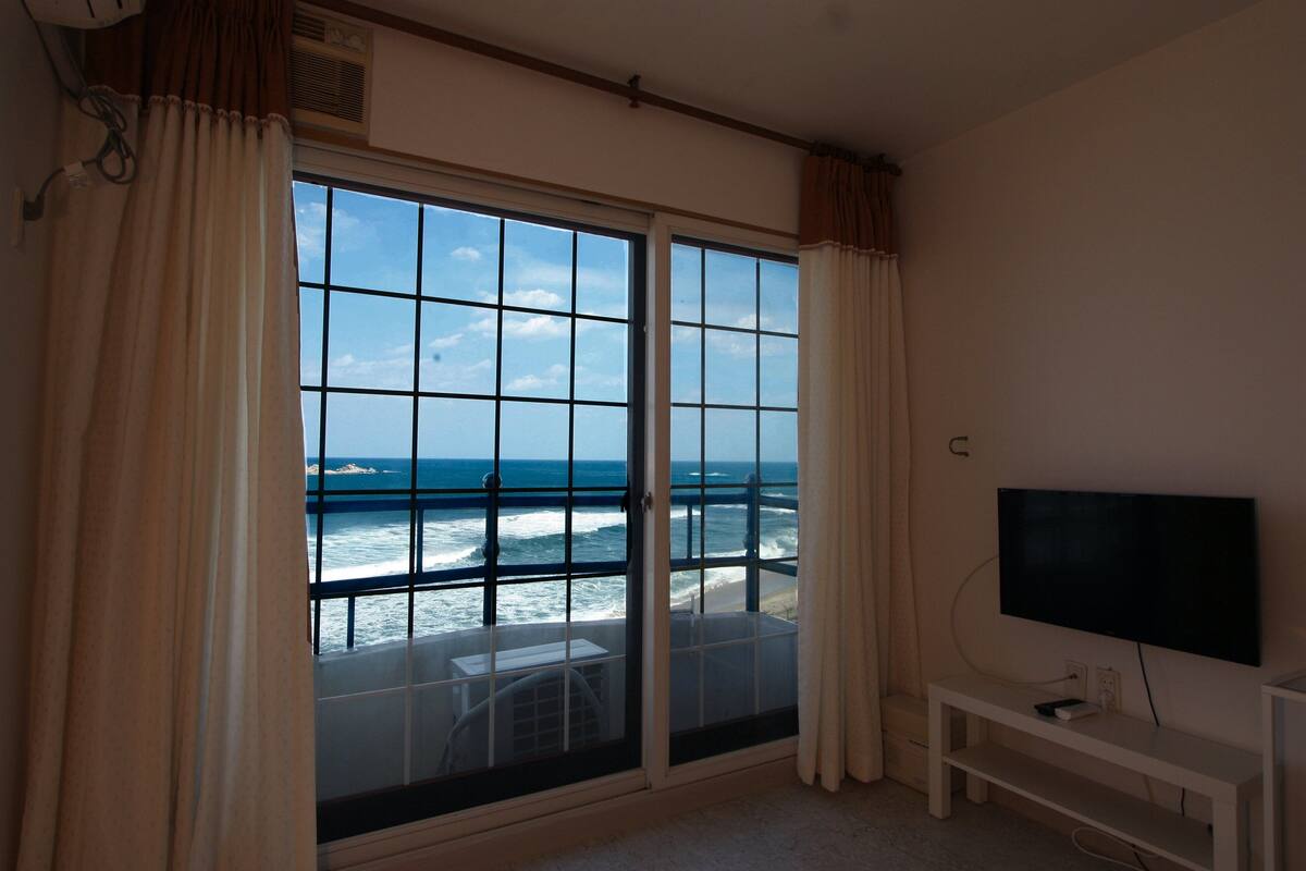 Een kamer met een groot raam met uitzicht op de oceaan, een televisie en een klein witte bankje.
