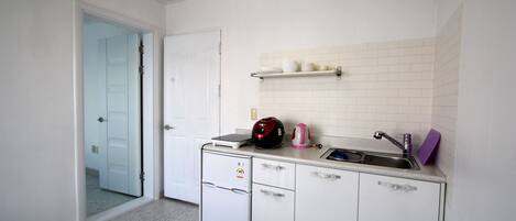 Une cuisine compacte avec des meubles blancs, un four encastré, un petit réfrigérateur, un évier et un plan de travail sur lequel se trouvent une bouilloire et des assiettes.