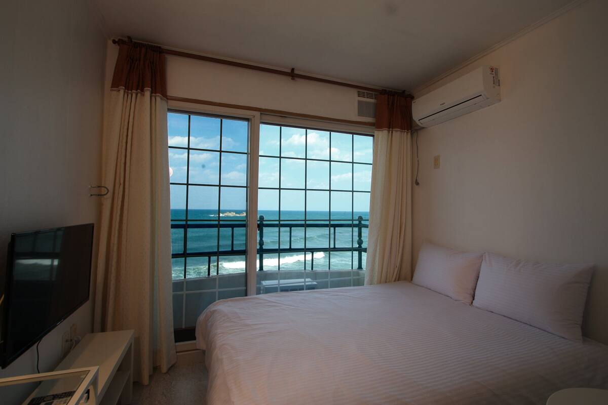 Een hotelkamer met een groot raam dat uitzicht biedt op de oceaan, een bed, een televisie en een airconditioner.