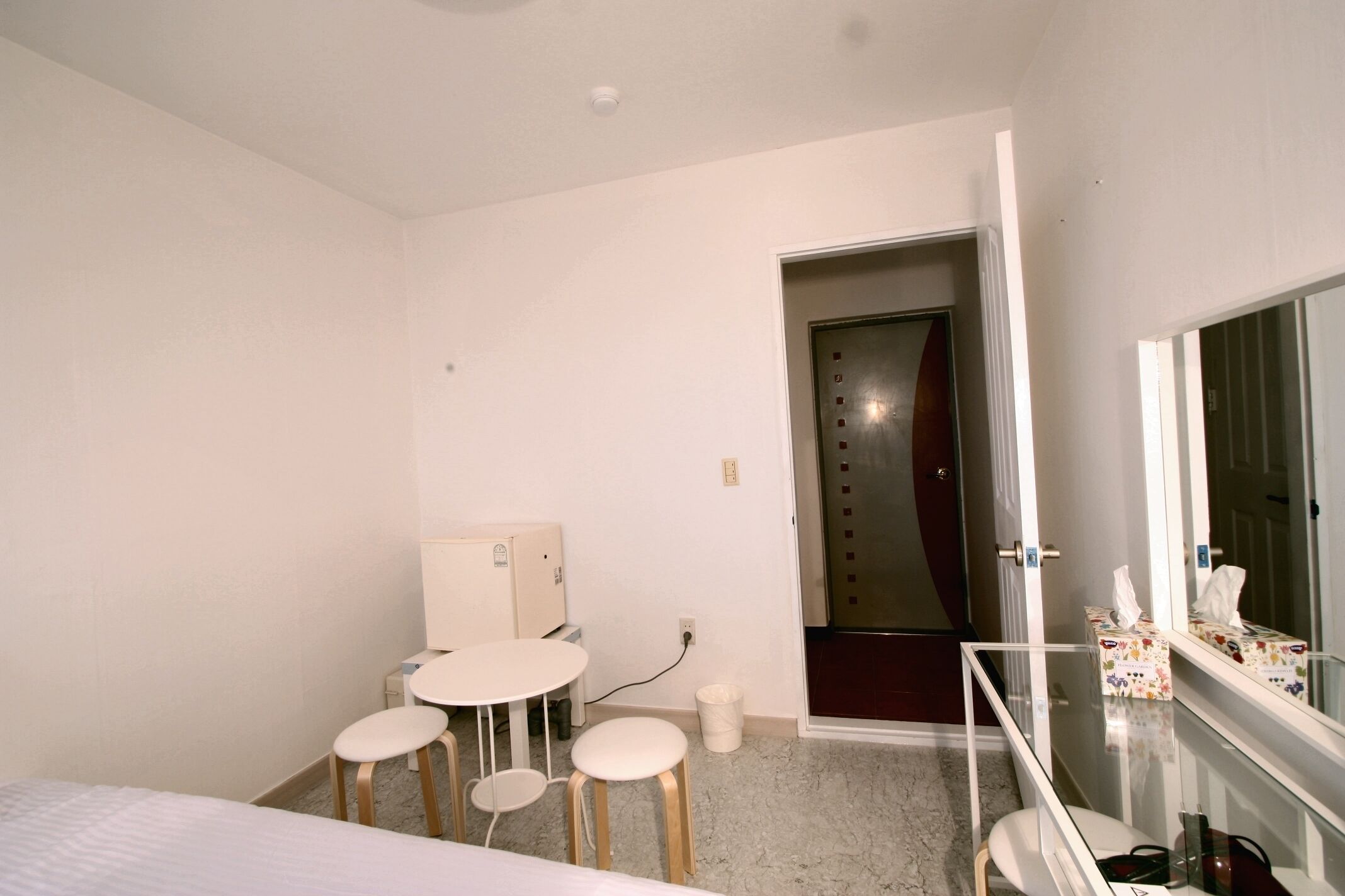 A small, modern hotel room with a bed, a round table with stools, a mirror, and a door leading to a hallway.