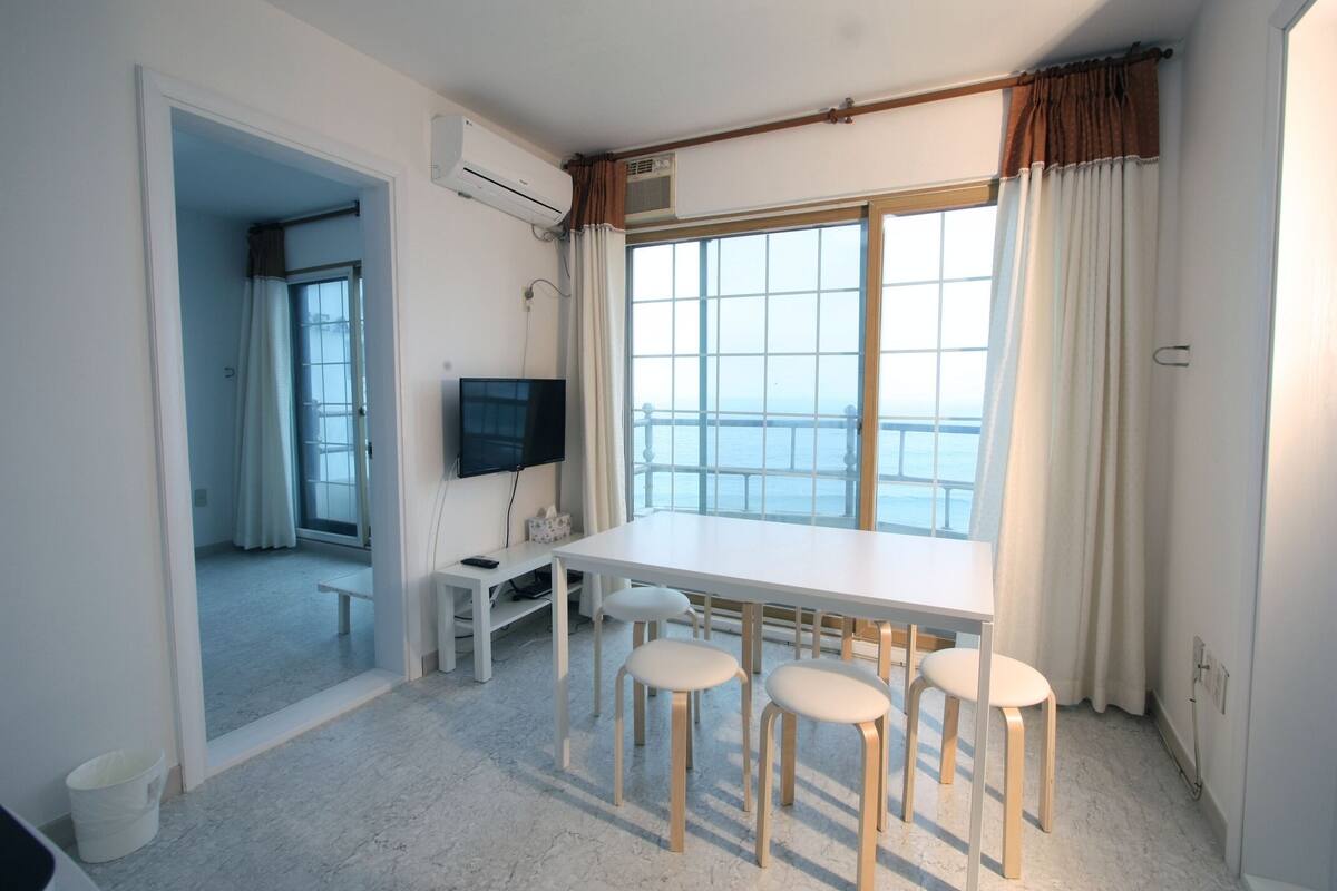 Een kamer met een groot raam, een flatscreen televisie, een witte tafel en vier witte stoelen.
