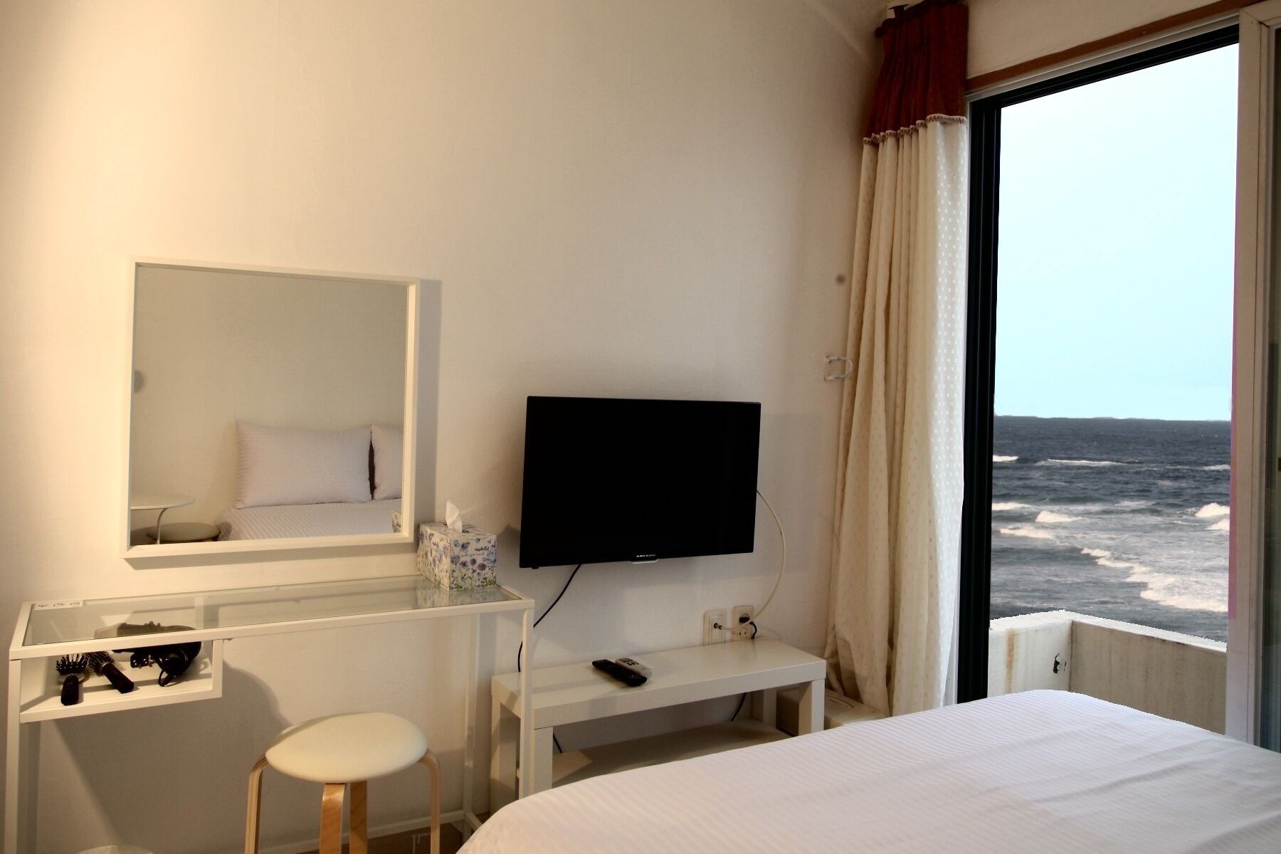 A hotel room with a bed, a TV, a desk with a chair, a mirror, and a view of the sea.