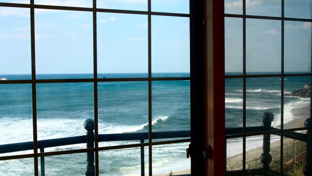 Vista a través de una ventana con diseño de cuadrícula, que da a una playa con olas y un faro distante.