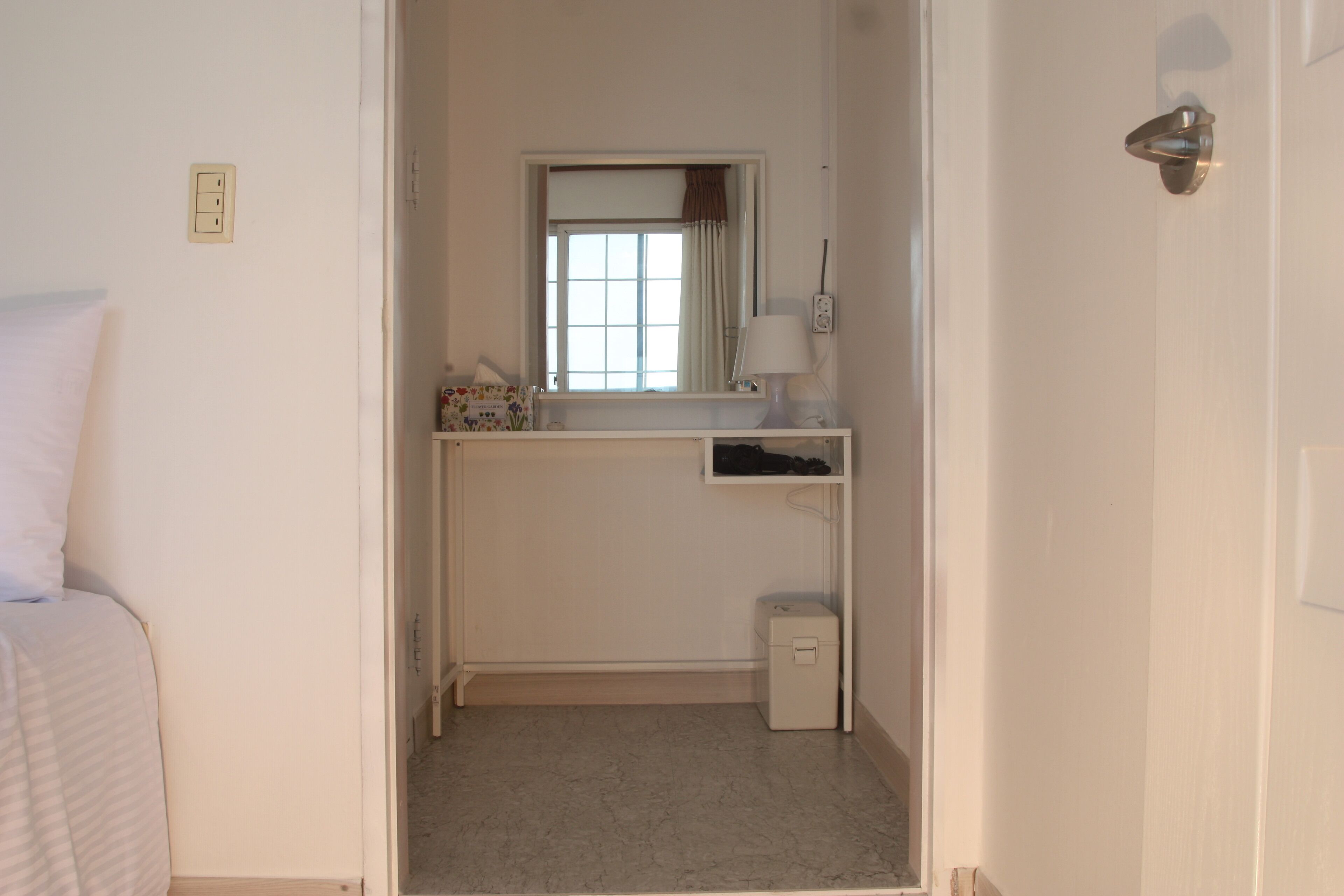 Ein kleiner, sauberer Raum mit einem Bett, einem Fenster mit Vorhängen, einem Spiegel, einem kleinen Tisch mit Lampe und einem Mülleimer.