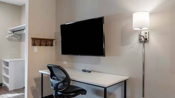 A hotel room with a flat-screen TV mounted on the wall, a desk with a chair, a coat rack, and a wall-mounted lamp.