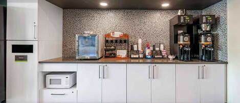 Uma cozinha moderna com armários brancos, micro-ondas, lixeira, espaço para café e painel decorativo.