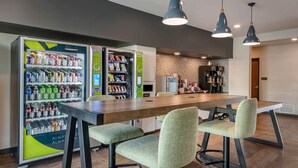 A modern dining area with a long wooden table, green upholstered chairs, and two vending machines stocked with snacks and drinks.