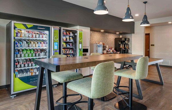 Un área de comedor moderna con una larga mesa de madera, sillas tapizadas en verde y dos máquinas expendedoras llenas de aperitivos y bebidas.
