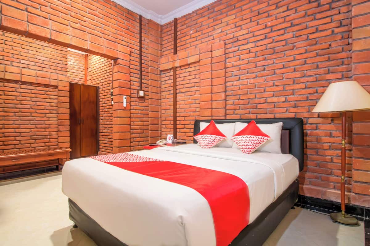A hotel room with a brick wall, a bed with a red and white bedspread, a nightstand, a lamp, and a wooden bench.