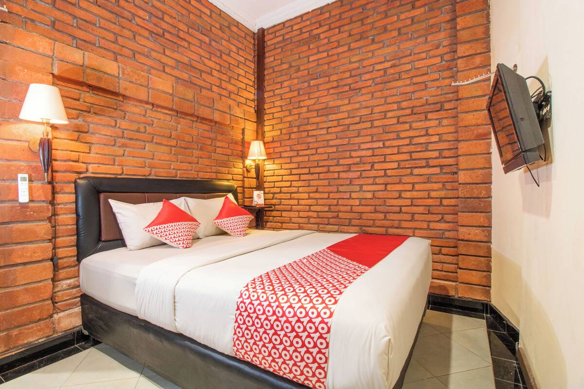 A hotel room with a brick wall, a bed with white bedding and red accents, two wall-mounted lamps, and a flat-screen TV.