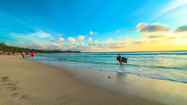 A beach with people walking and a surfer carrying a surfboard. - Hotel O Maxi Budget (Kuta)