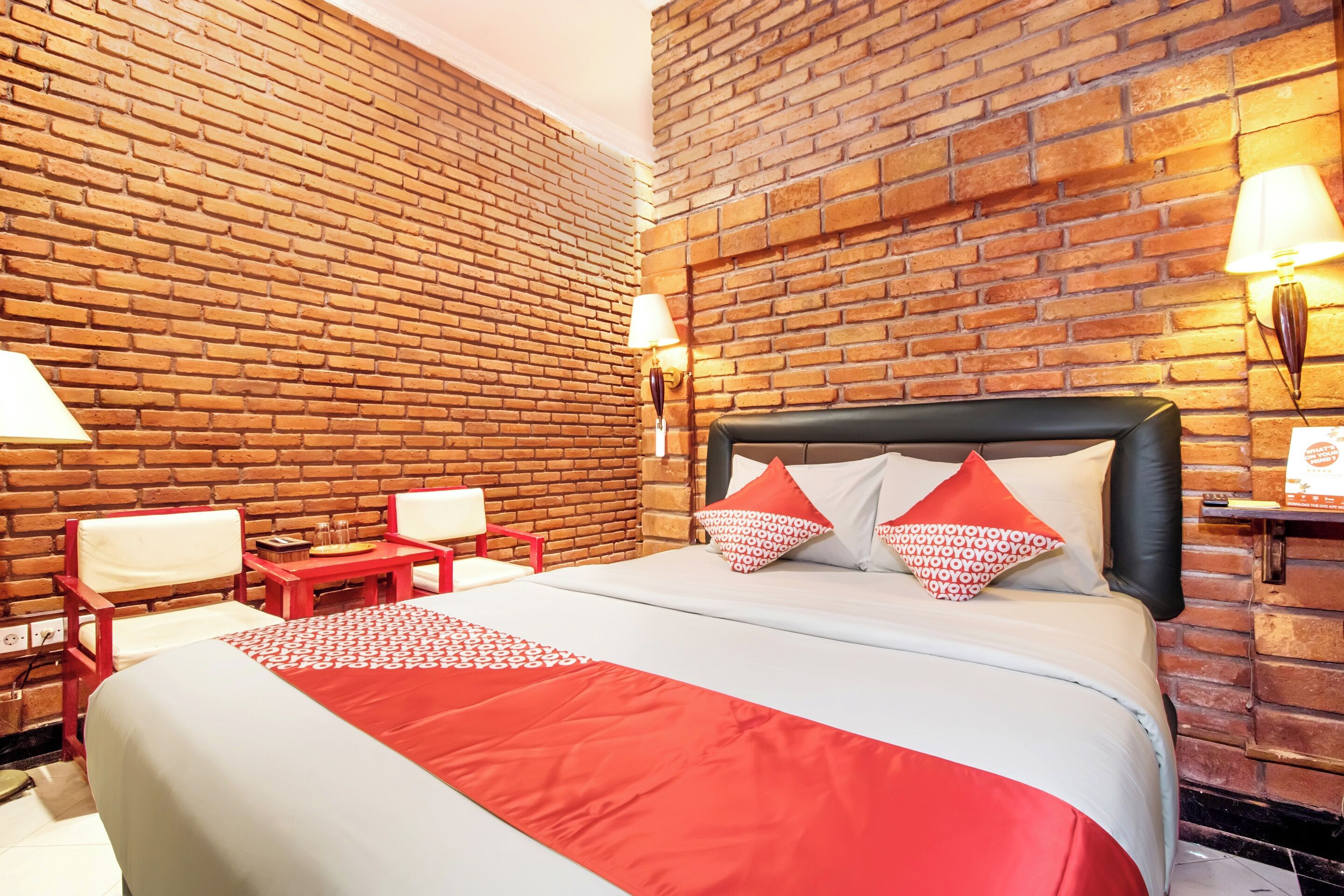 A hotel room with a brick wall, a bed with red and white bedding, two chairs, a small table, and two wall-mounted lamps.