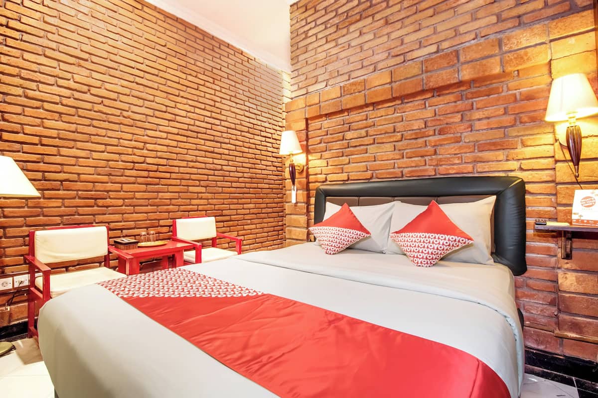 A hotel room with a brick wall, a bed with red and white bedding, two chairs, a small table, and two wall-mounted lamps.