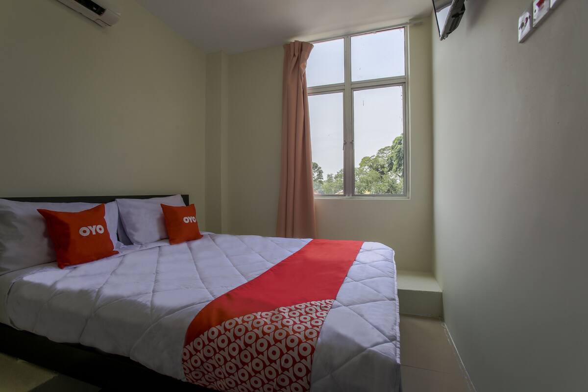 Ein ordentlich bezogenes Bett mit einer gemusterten Decke und zwei Kissen, ein Fenster mit Blick auf Bäume und eine Wand-Klimaanlage.