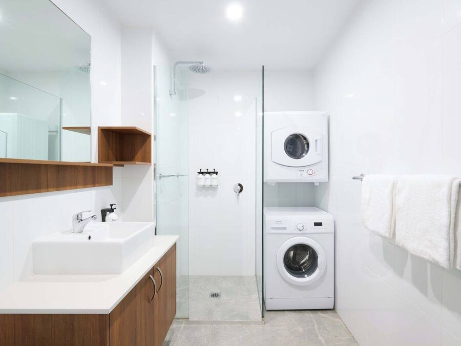 Un baño moderno con lavamanos, espejo, armarios de madera, cabina de ducha de vidrio y lavadora y secadora apiladas.