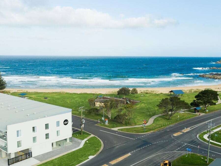 Une route côtière avec une plage en arrière-plan, un bâtiment moderne doté d’un toit plat et un ciel dégagé.