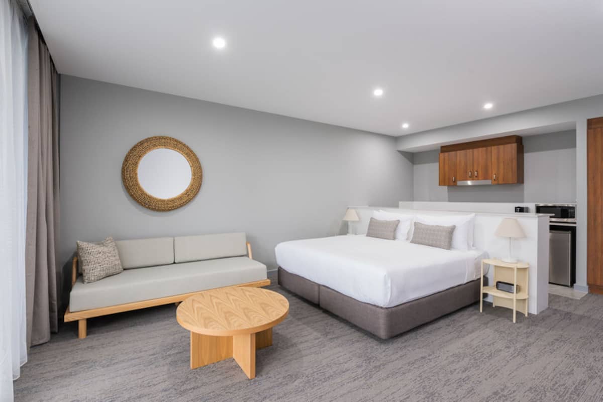 Une chambre d’hôtel moderne avec un canapé gris, un grand lit, une table basse en bois, un miroir rond et un coin cuisine.