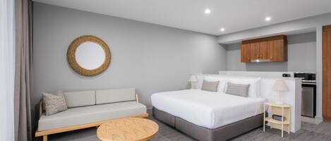 Une chambre d’hôtel moderne avec un canapé gris, un grand lit, une table basse en bois, un miroir rond et un coin cuisine.