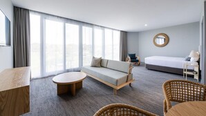 A modern hotel room with a sofa, a round coffee table, wicker chairs, a bed, and a large window offering a city view. - Abode Hotel Malua Bay (Malua Bay)