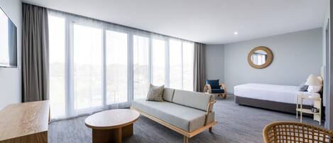 A modern hotel room with a sofa, a round coffee table, wicker chairs, a bed, and a large window offering a city view.