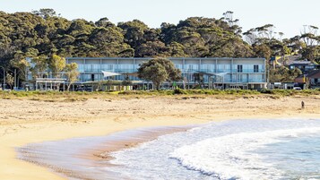 Un hotel frente a la playa con una playa arenosa, olas del océano y una colina boscosa al fondo.