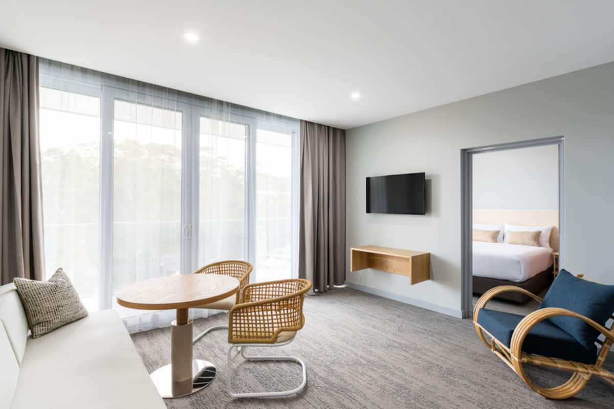 Une chambre d’hôtel moderne équipée d’un canapé, d’une table de salle à manger ronde avec des chaises, d’un téléviseur à écran plat fixé au mur, d’un lit avec une tête de lit et de grandes fenêtres avec des rideaux.