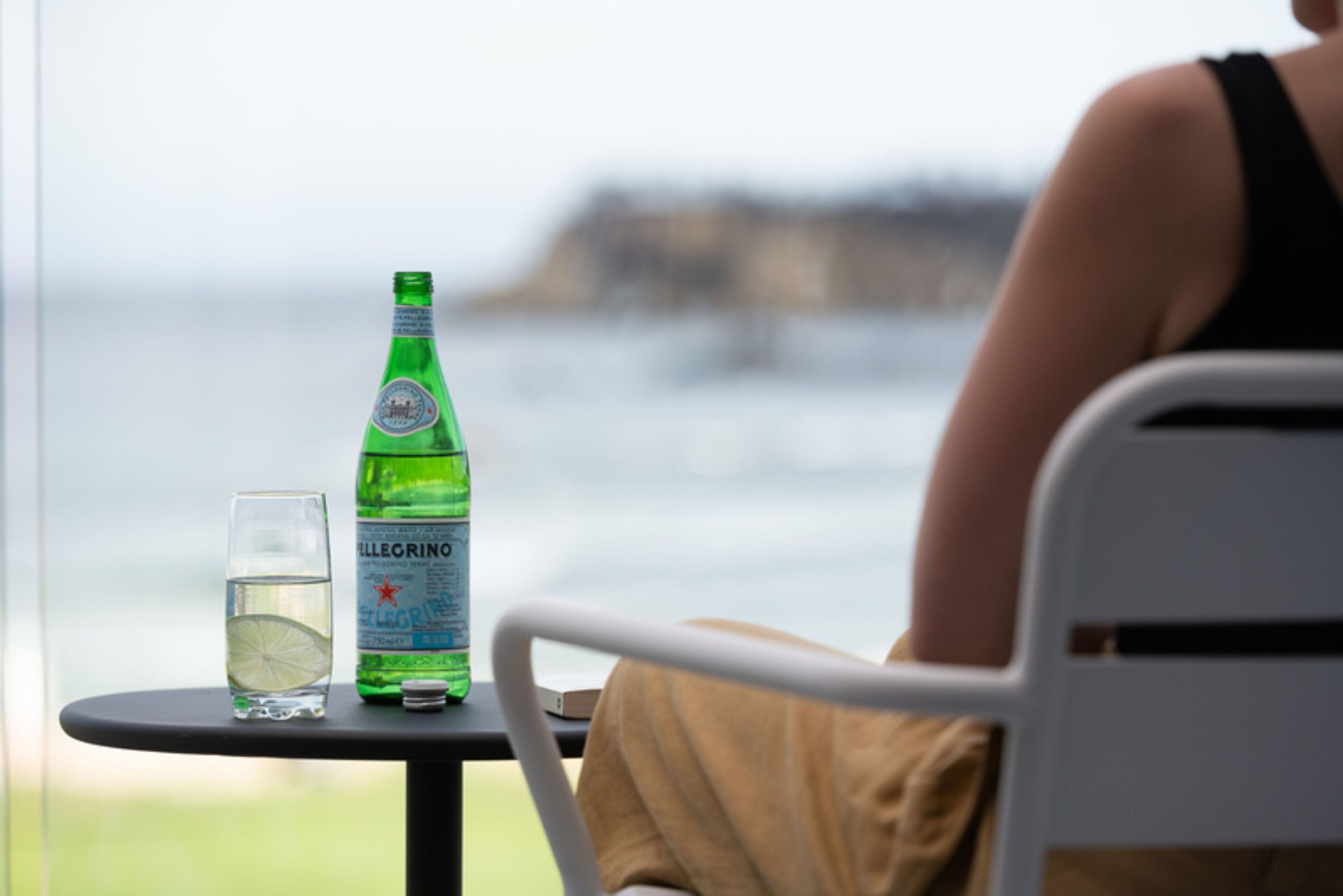 Une table avec une bouteille d’eau Pellegrino et un verre, une personne assise à la table, et une vue sur la mer.
