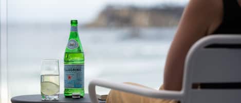 A table with a bottle of Pellegrino water and a glass, a person seated at the table, and a view of the sea.