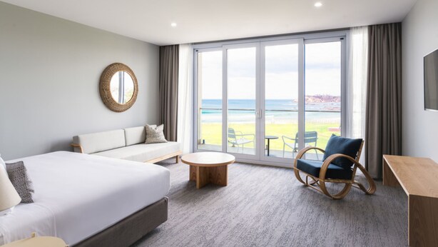 A modern hotel room with a large bed, a sofa, a round coffee table, and a wicker chair. The room features a large window with a view of the beach and ocean. - Abode Hotel Malua Bay (Malua Bay)