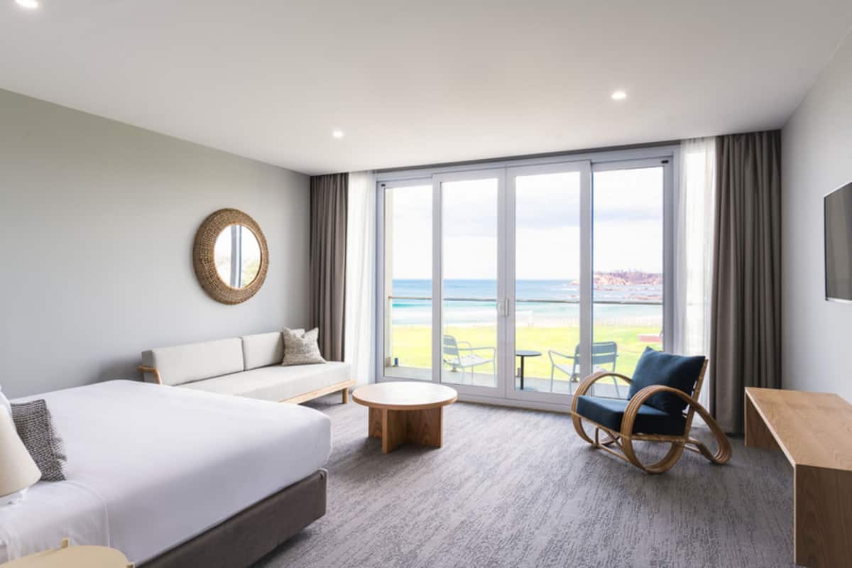 Une chambre d’hôtel moderne dotée d’un grand lit, d’un canapé, d’une table basse ronde et d’un fauteuil en osier. La chambre dispose d’une grande fenêtre offrant une vue sur la plage et l’océan.