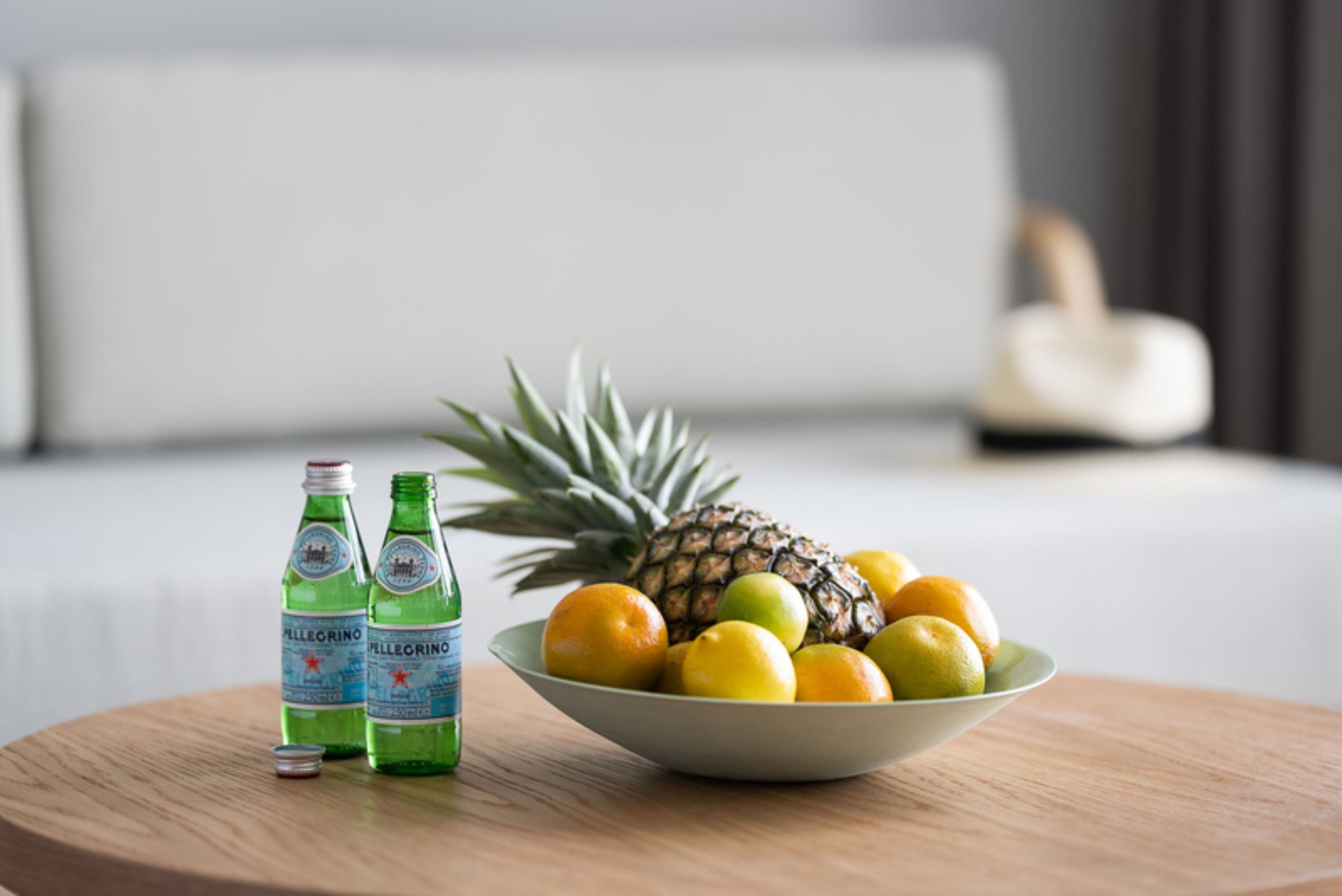 A bowl of fruits and Pellegrino water bottles on a wooden table in a hotel room.