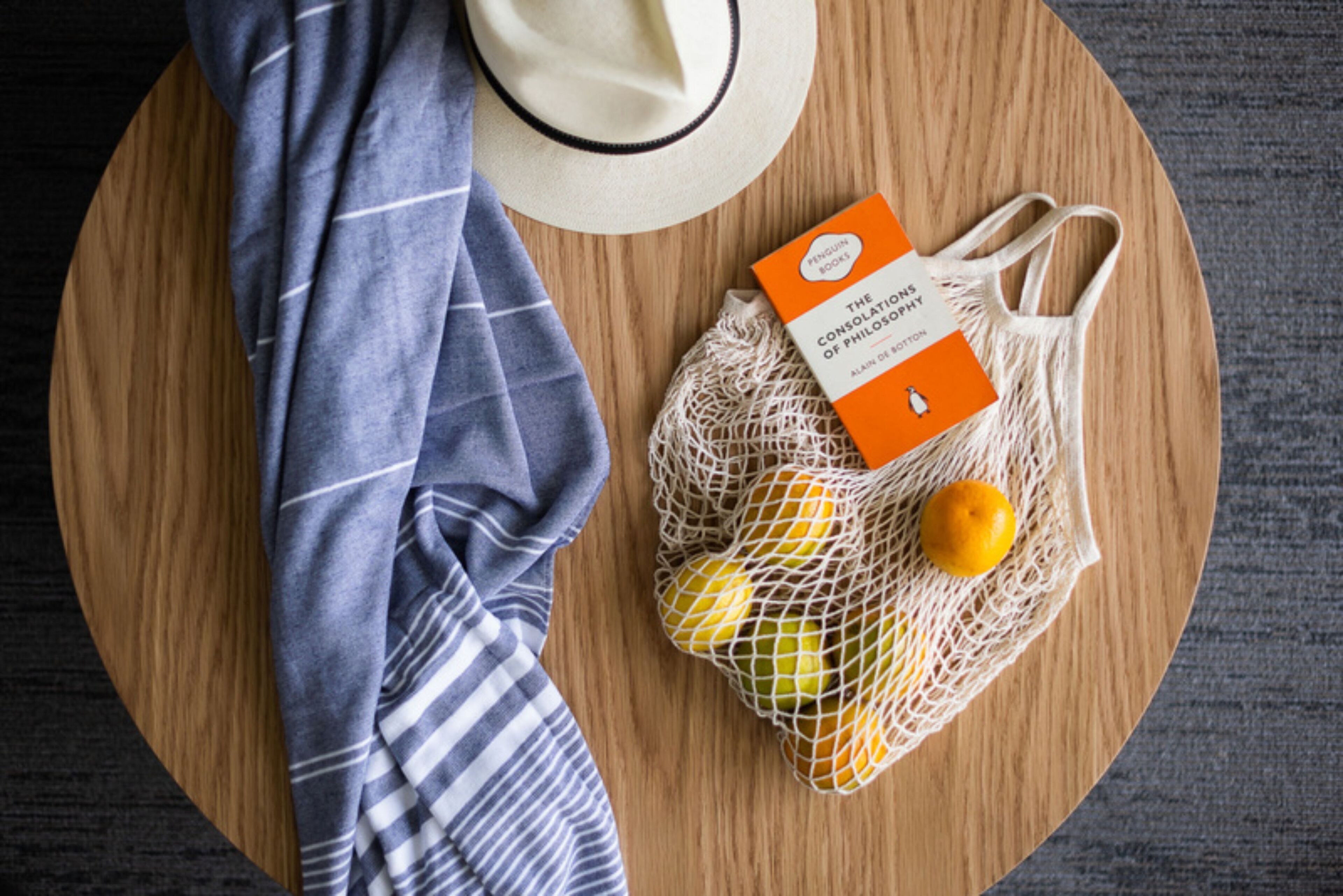 A round wooden table with a white hat, a striped cloth, and a mesh bag containing oranges and a book titled "The Consolations of Philosophy" by Alain De Botton.