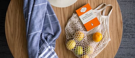A round wooden table with a white hat, a striped cloth, and a mesh bag containing oranges and a book titled "The Consolations of Philosophy" by Alain De Botton.