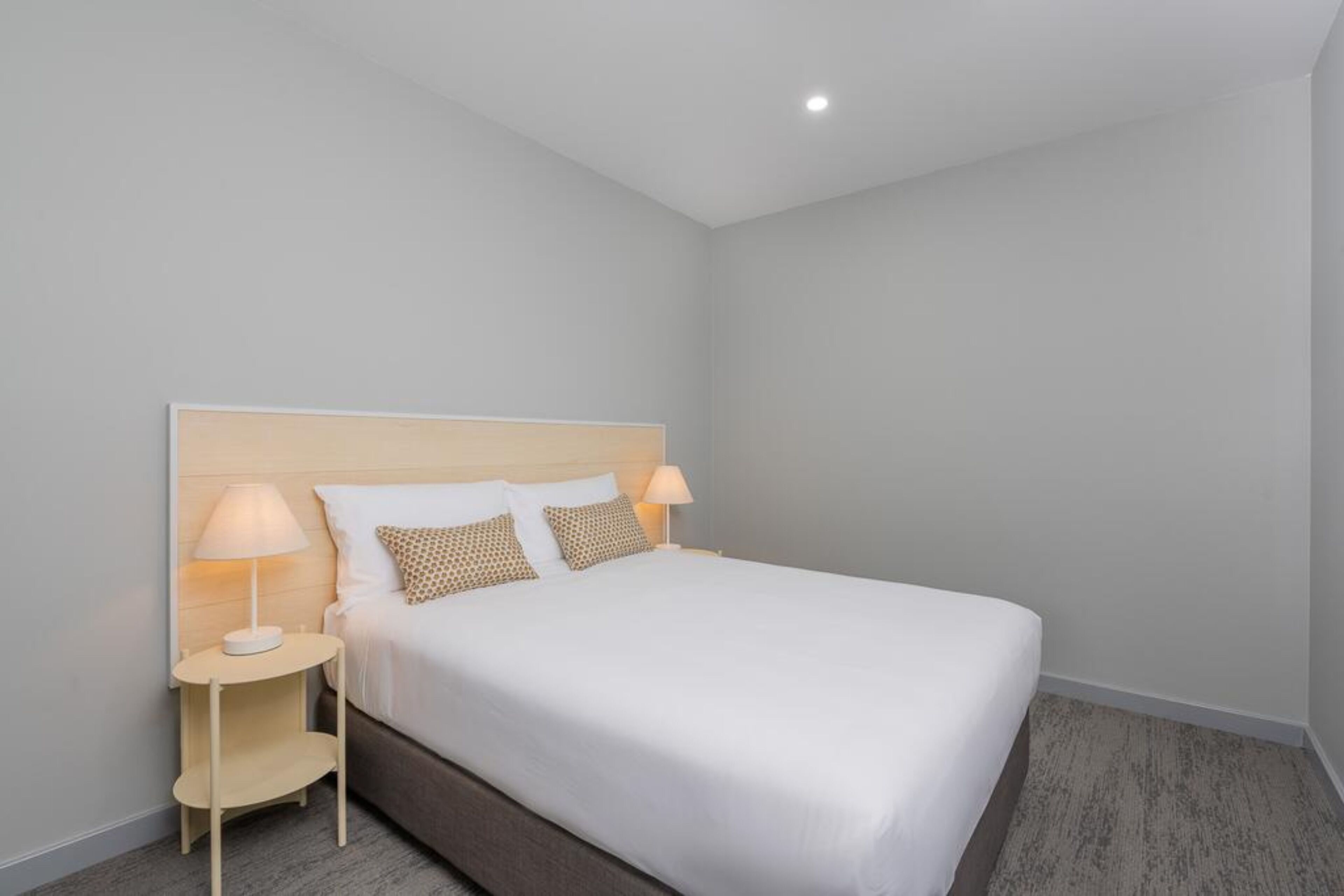 A neatly made bed with a wooden headboard, two bedside tables with lamps, and a light-colored carpet.