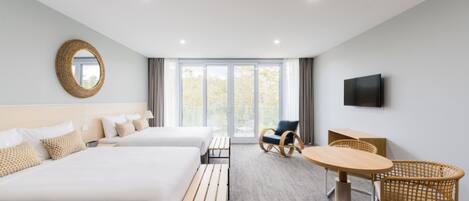 A hotel room with two beds, a wooden bench, a round table, wicker chairs, a television, and large windows.
