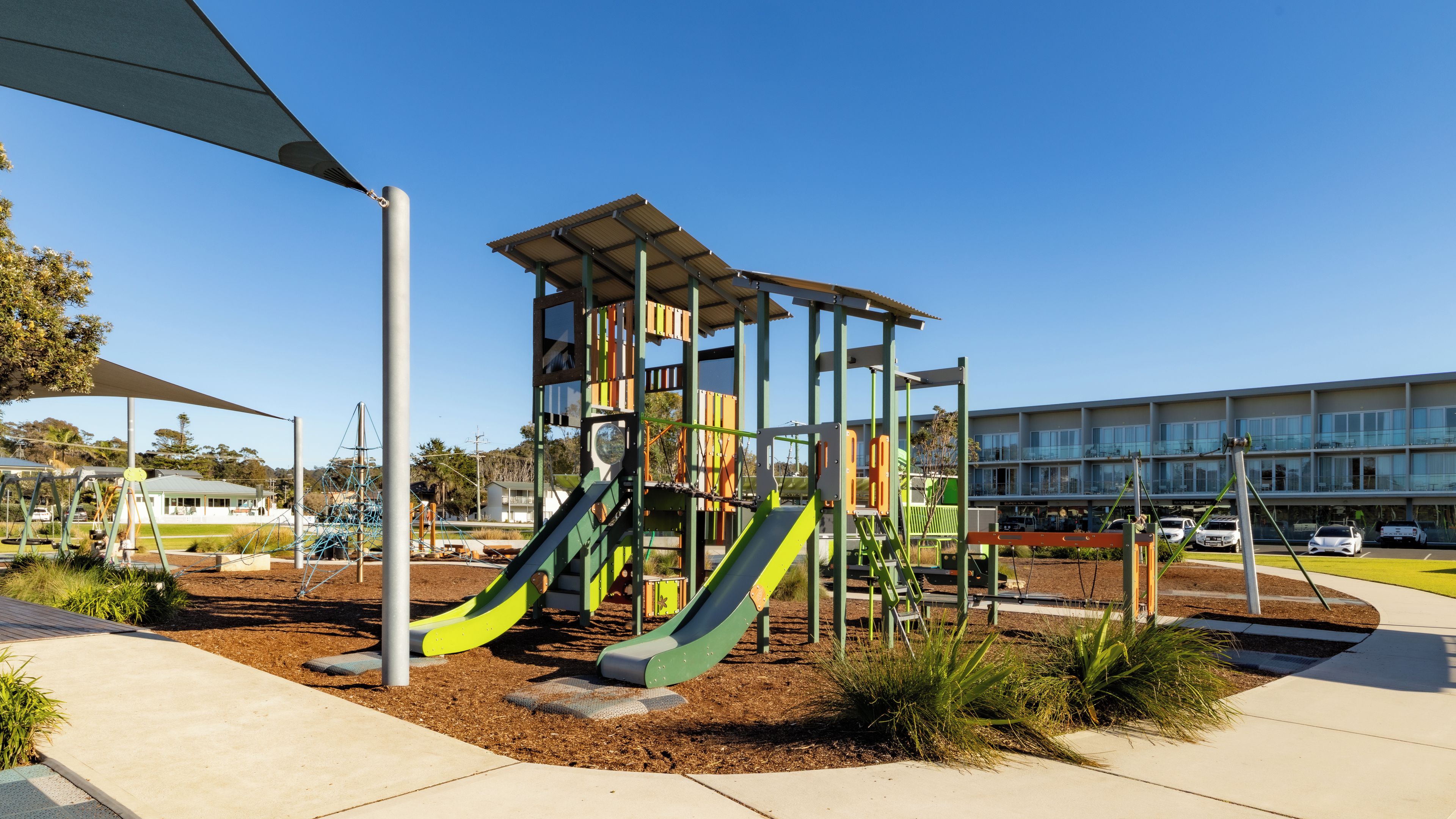 A playground with slides and climbing structures, surrounded by a paved area and a building in the background.