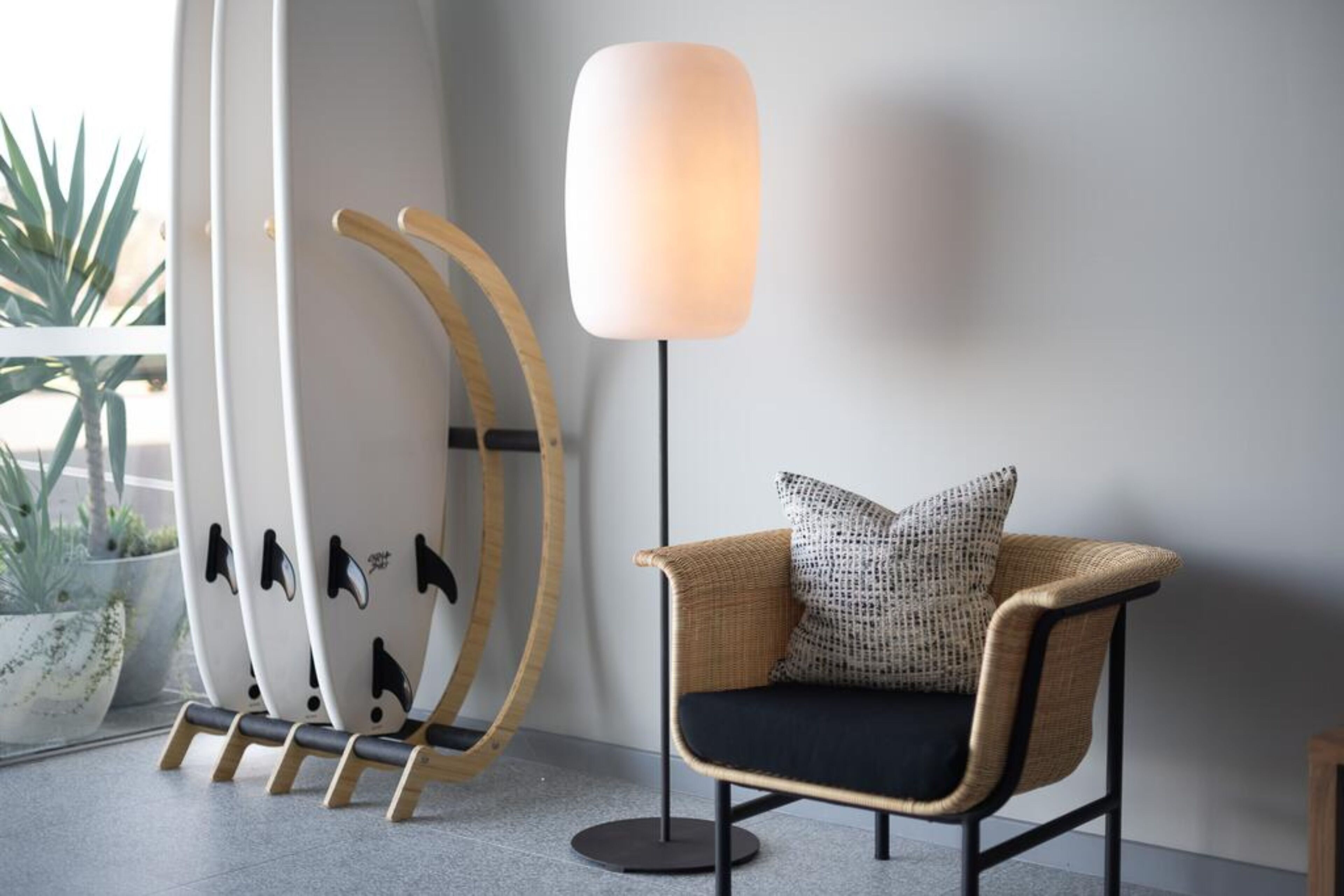 A modern interior with a wicker chair, a floor lamp, and surfboards leaning against a wall.