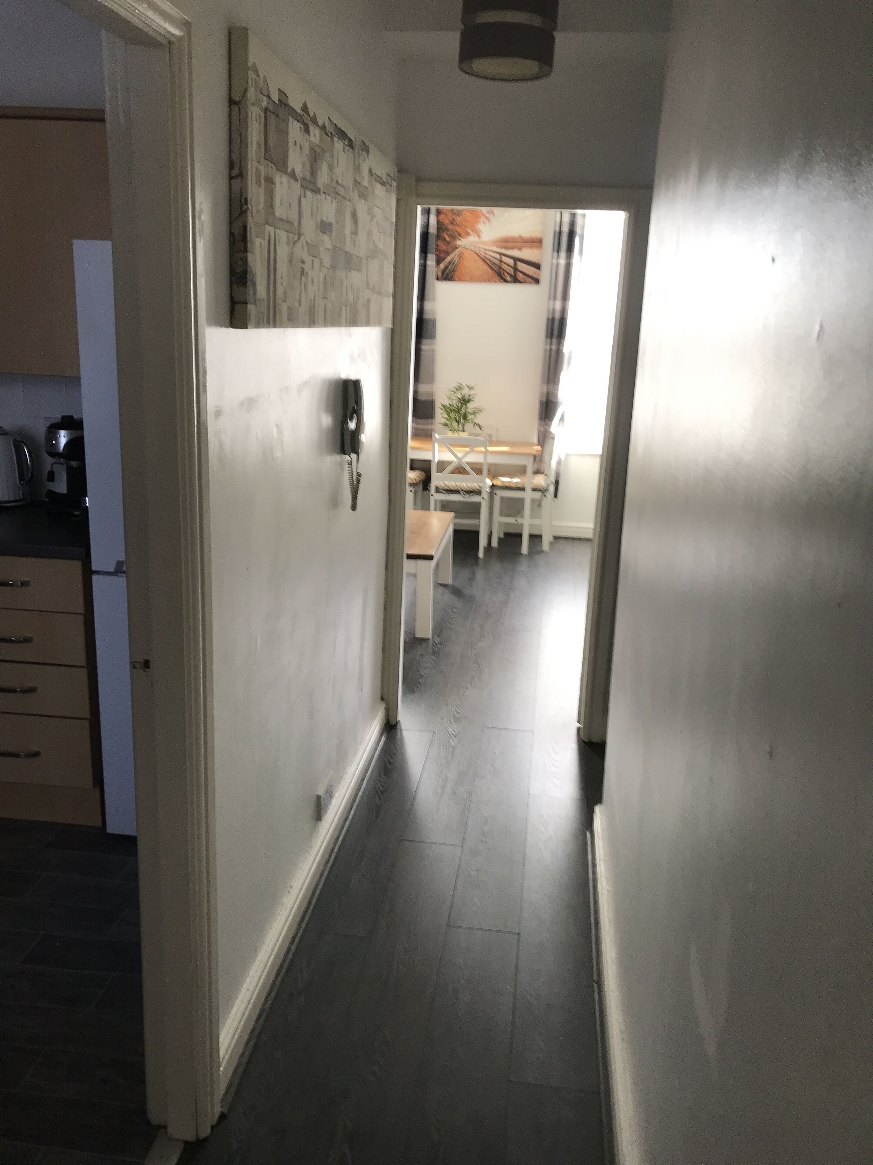 Un couloir étroit, avec un coin cuisine sur la gauche, une table à manger avec des chaises et, au fond, une fenêtre avec des rideaux.