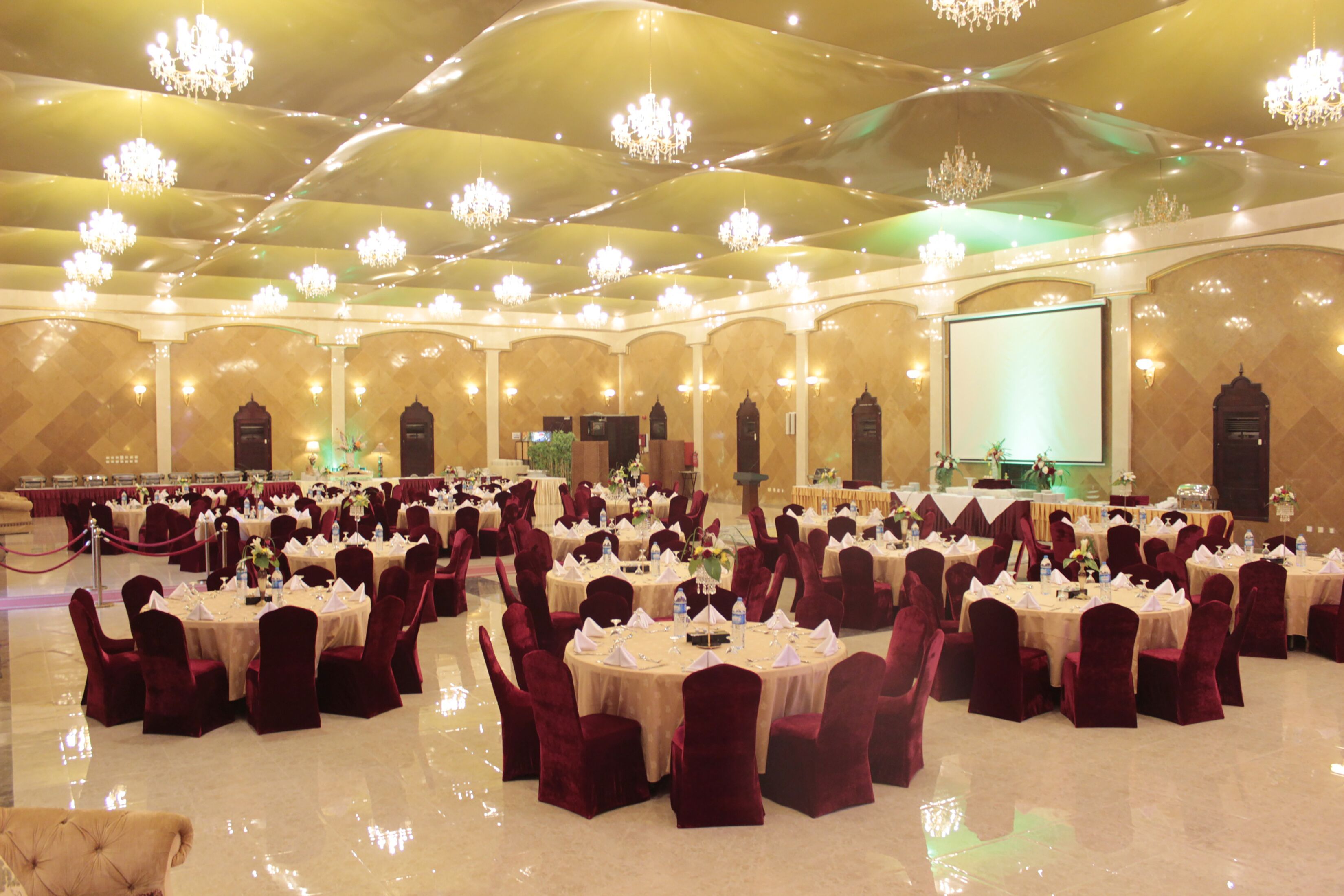 A banquet hall with round tables, red velvet chairs, chandeliers, and a stage.