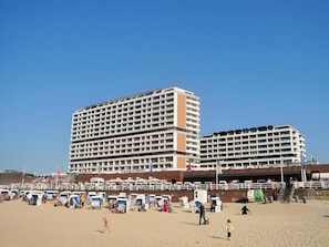 Um hotel grande com vários andares e varandas, uma praia de areia em frente, e cadeiras e guarda-sóis de praia.