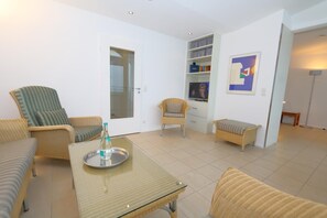 Uma sala de estar com mesa de vidro, móveis de vime, televisão e estante de livros.