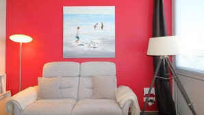 Une pièce avec un mur rouge, un canapé gris, un tableau représentant des enfants qui jouent, un lampadaire et une fenêtre avec des stores.