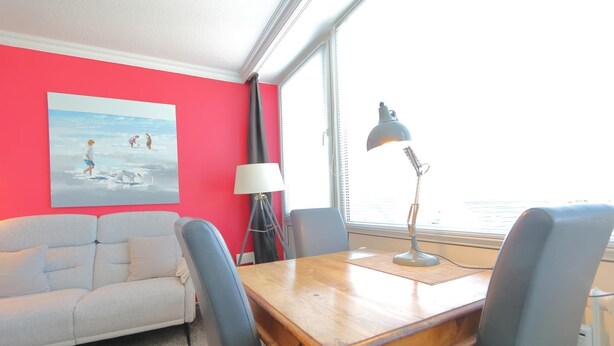 Une pièce avec un mur rouge, un canapé blanc, une table en bois avec des chaises, un lampadaire et une fenêtre donnant sur un paysage.
