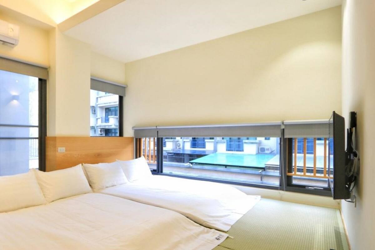 Una habitación de hotel con cama, ventana y vistas a edificios, además de un equipo de aire acondicionado.
