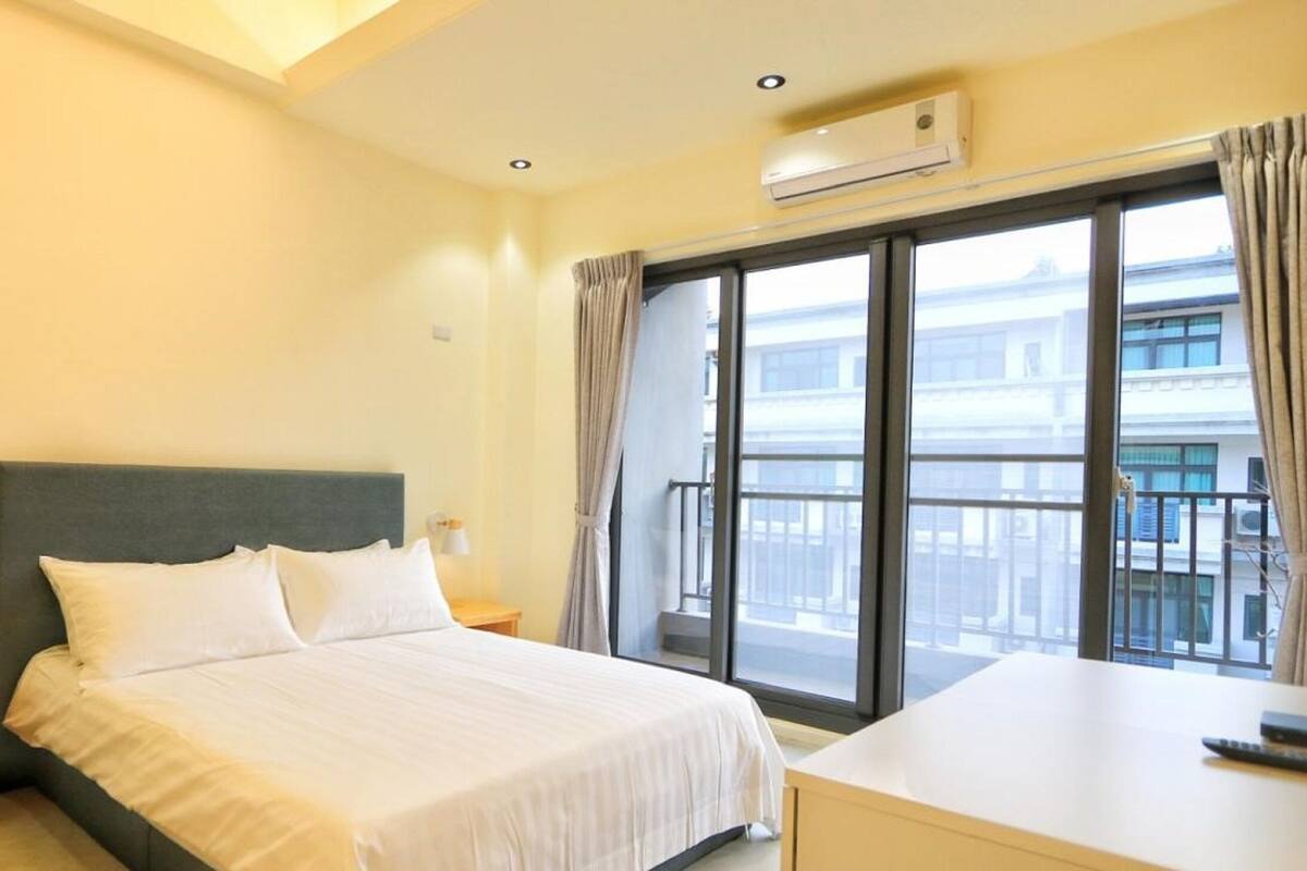 Habitación de hotel con cama, mesitas de noche, escritorio y un ventanal con vistas a edificios.