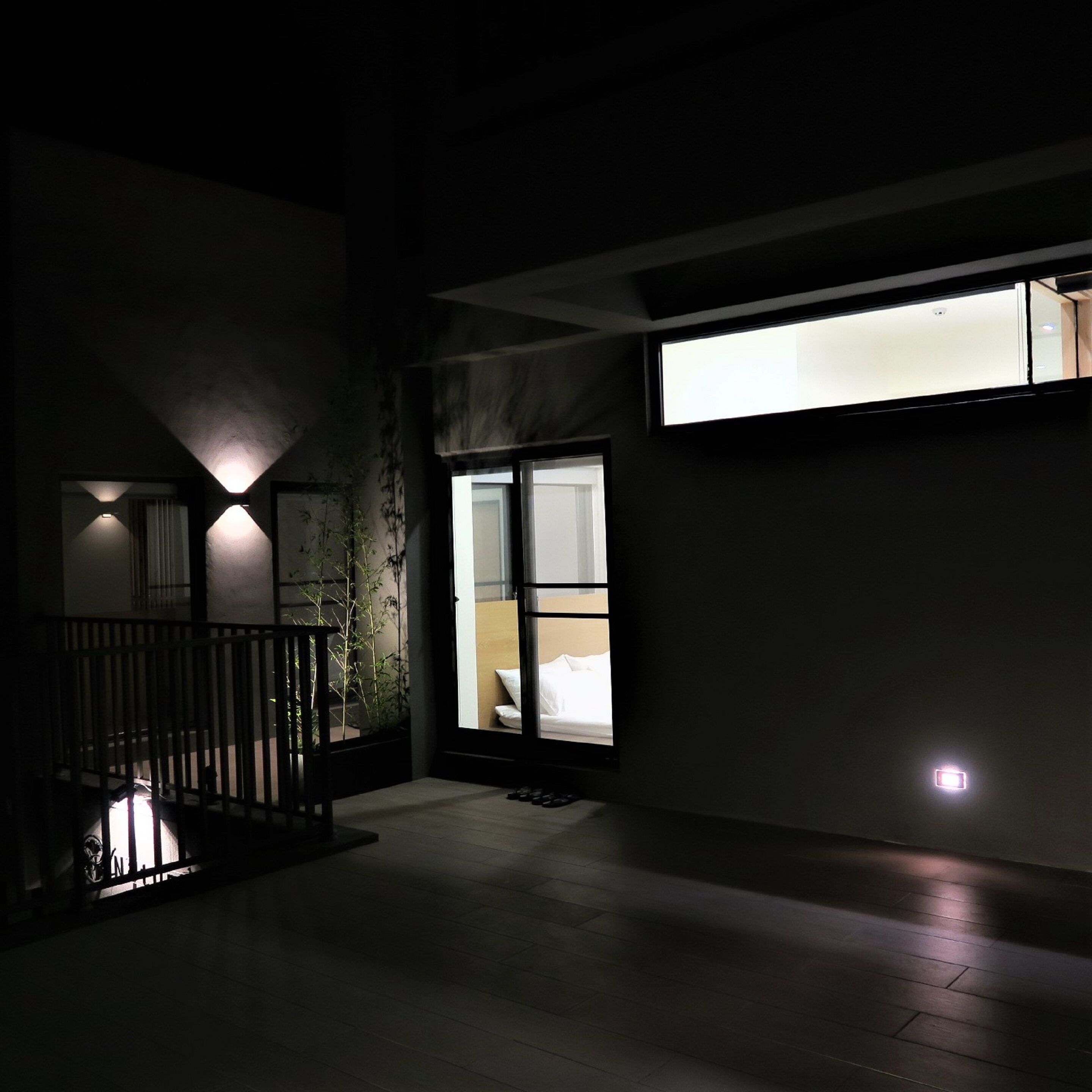 A dimly lit outdoor area with a staircase leading to a balcony, a door with a view into a room, and a small illuminated light fixture on the ground.